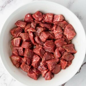 A white bowl filled with raw, cubed steak bites marinated with seasoning, placed on a white marble surface.