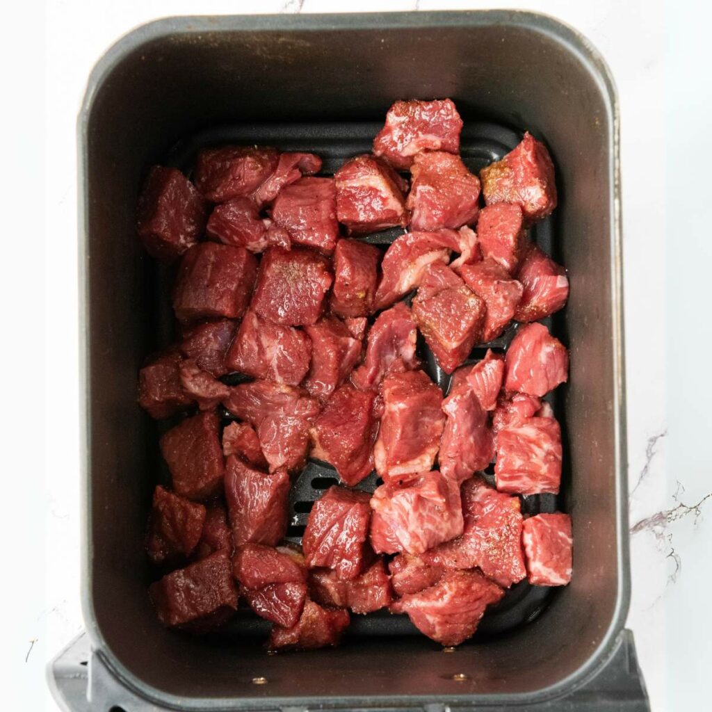 Raw cubed beef pieces arranged in a single layer at the bottom of an air fryer basket.