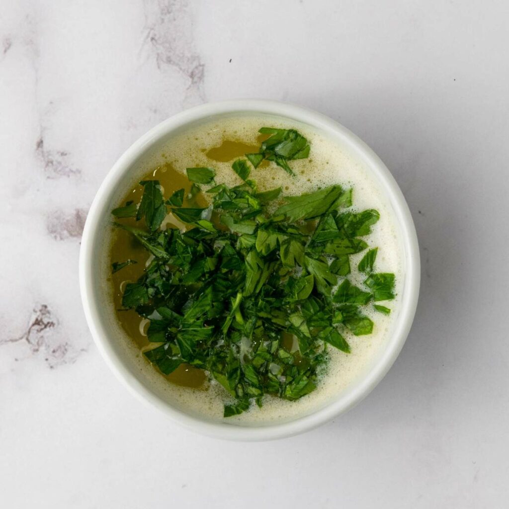 A white bowl filled with melted butter topped with chopped fresh parsley.