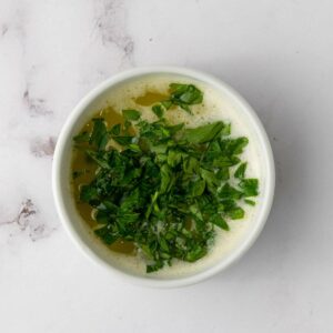 A white bowl filled with melted butter topped with chopped fresh parsley on a marble surface.