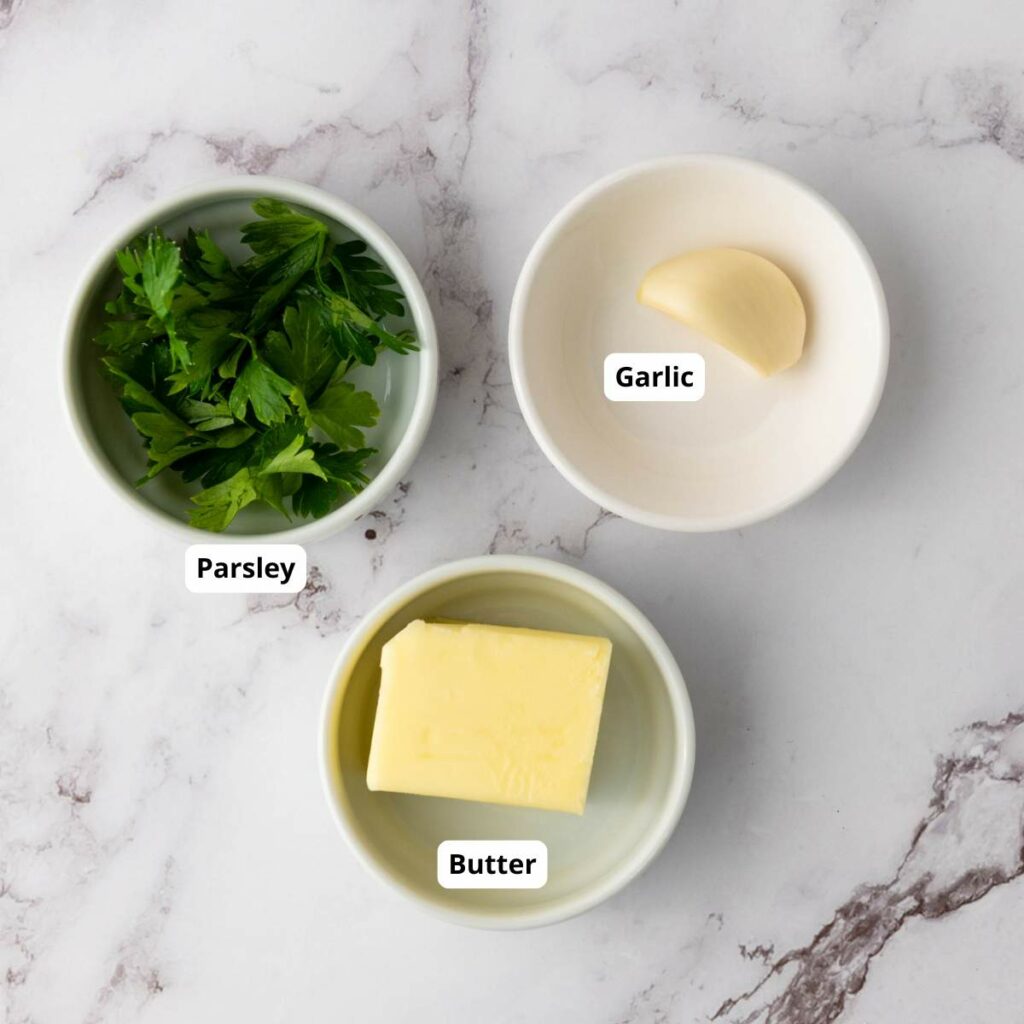 Three small bowls containing parsley, a clove of garlic, and a piece of butter.