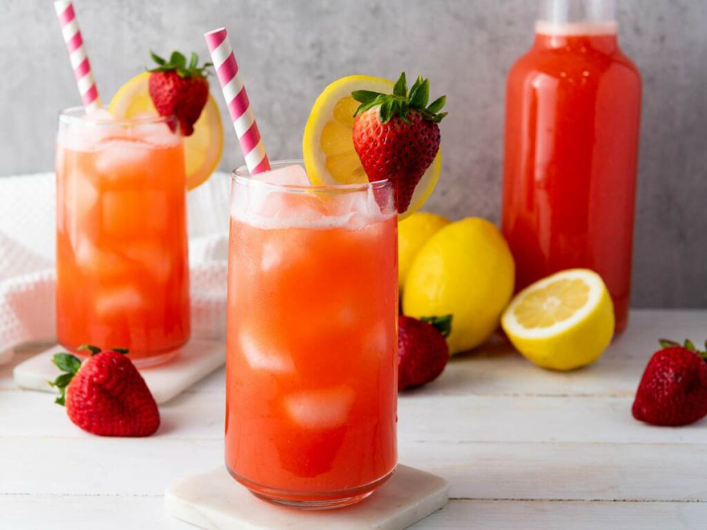Two glasses of iced strawberry lemonade with striped straws, garnished with lemon slices and strawberries.