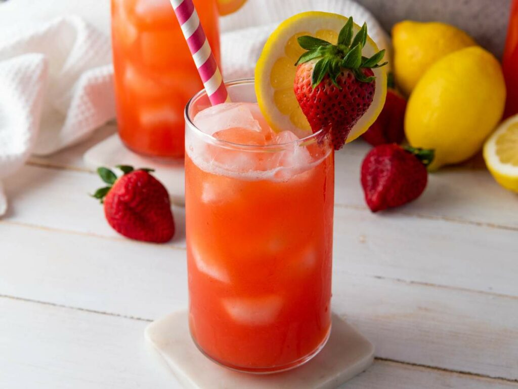 A glass of strawberry lemonade garnished with a lemon slice and a whole strawberry, with extra strawberries and lemons in the background.
