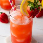 A glass of iced strawberry lemonade garnished with a lemon slice and a strawberry, with a pink-striped straw on a white coaster.