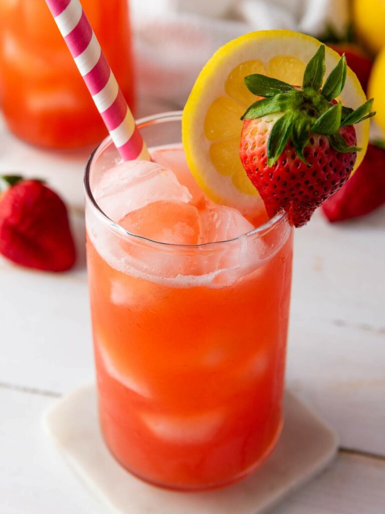 A glass of iced strawberry lemonade garnished with a lemon slice and a strawberry, with a pink-striped straw on a white coaster.