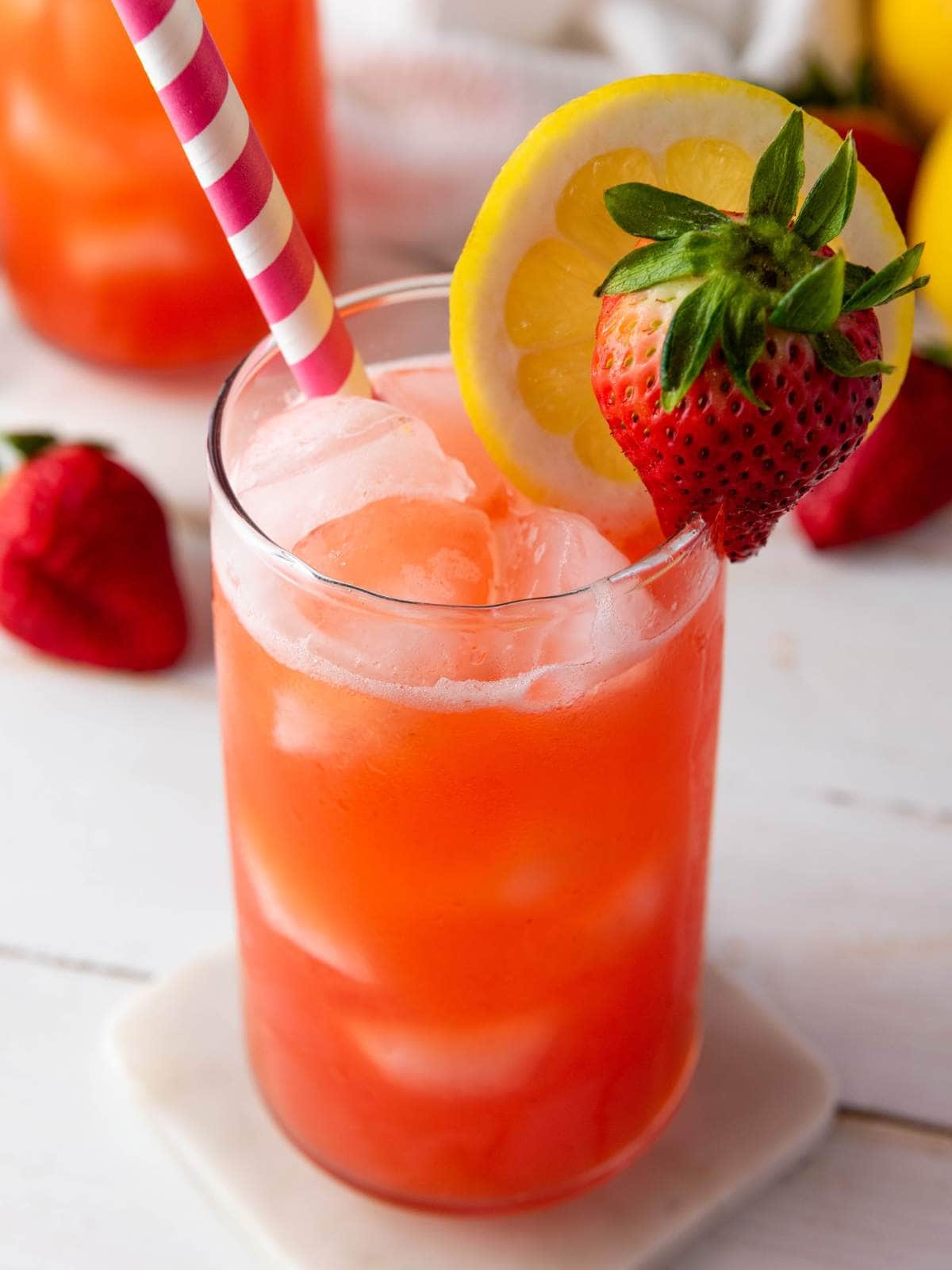 A glass of iced strawberry lemonade garnished with a lemon slice and a strawberry, with a pink-striped straw on a white coaster.