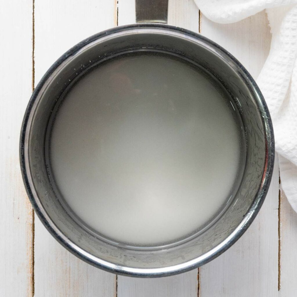 A stainless steel pot filled with syrup sits on a white wooden surface next to a white textured cloth.