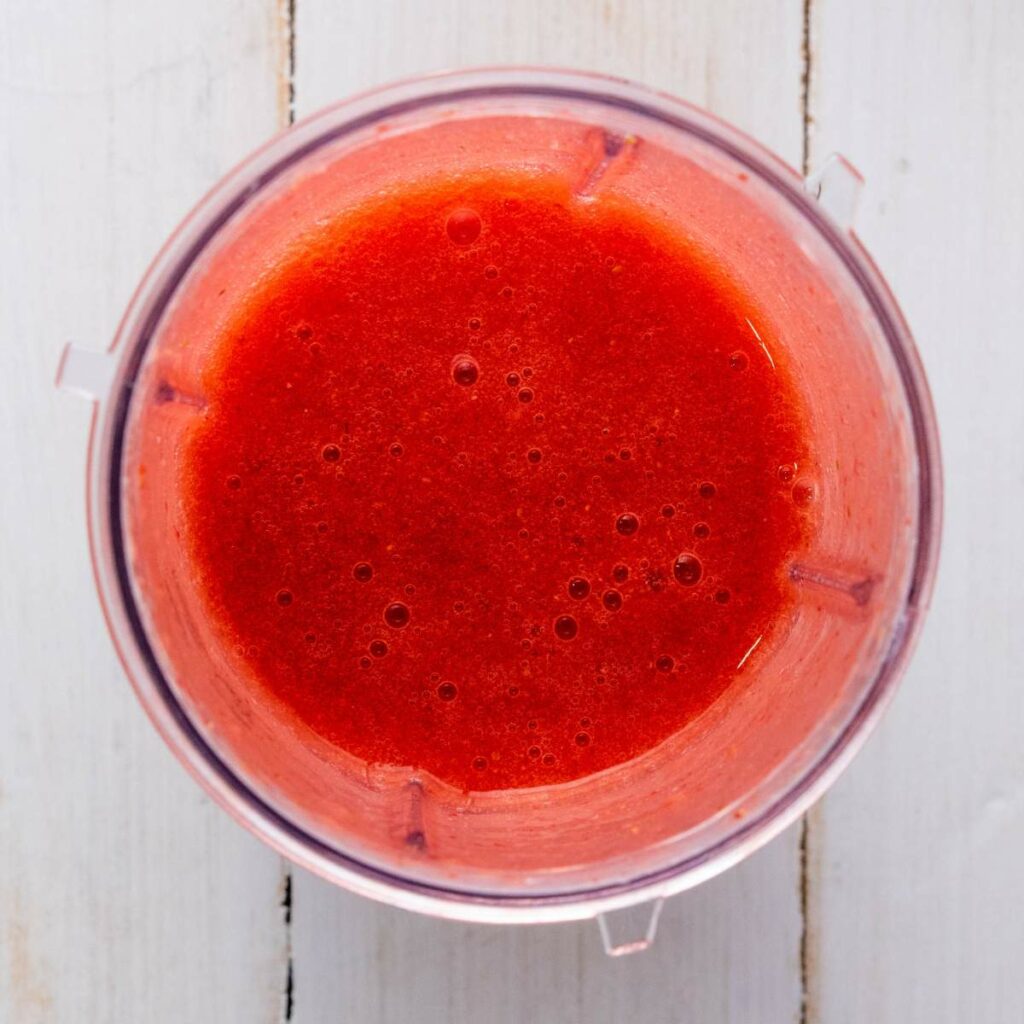 A top-down view of a blender filled with strawberry puree on a white wooden surface.