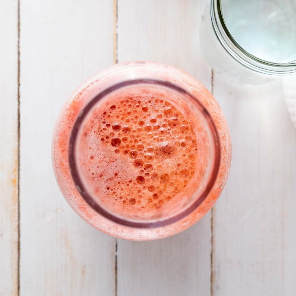Overhead view of a glass filled with a frothy pink beverage on a light wooden surface.