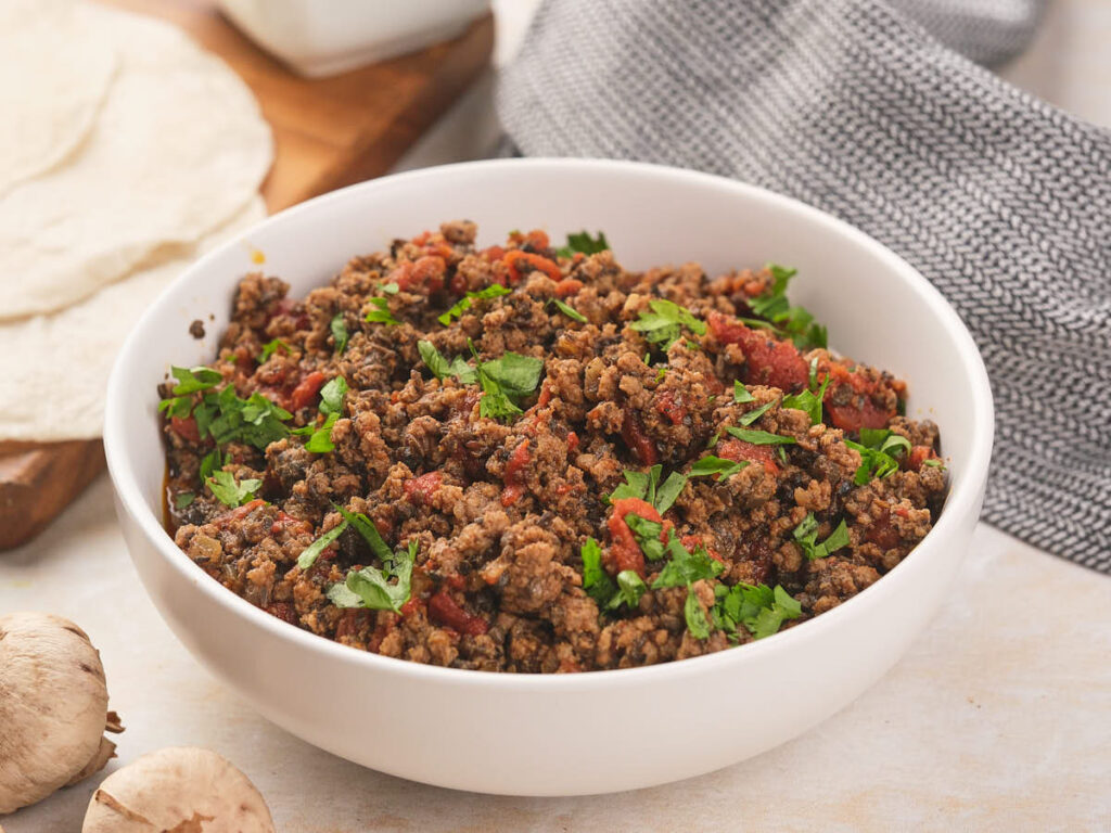 A white bowl of taco meat garnished with fresh cilantro alongside with two garlic cloves, tortillas and a table cloth.