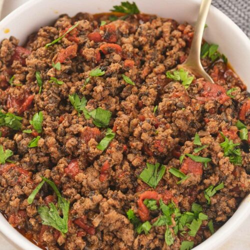 A white bowl of cooked meat garnished with fresh cilantro with a spoon inside.