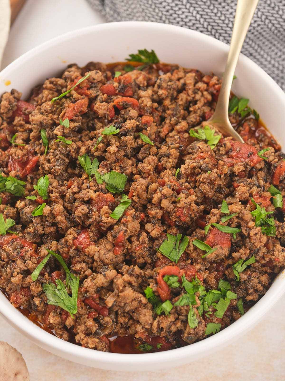 A white bowl of cooked meat garnished with fresh cilantro with a spoon inside.