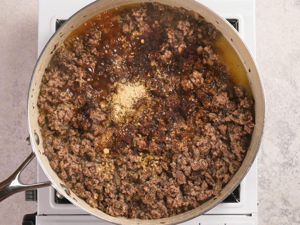 A simmering ground beef seasoned with spices in a white skillet on a stovetop.