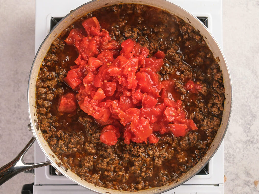 A skillet of cooked ground beef is being simmered with a diced tomatoes added on top.