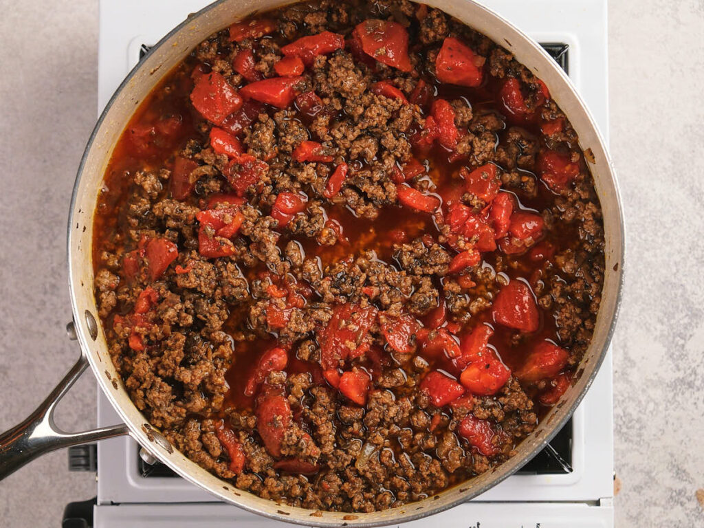 A seasoned ground beef and diced tomatoes are combined and being cooked in a white skillet.