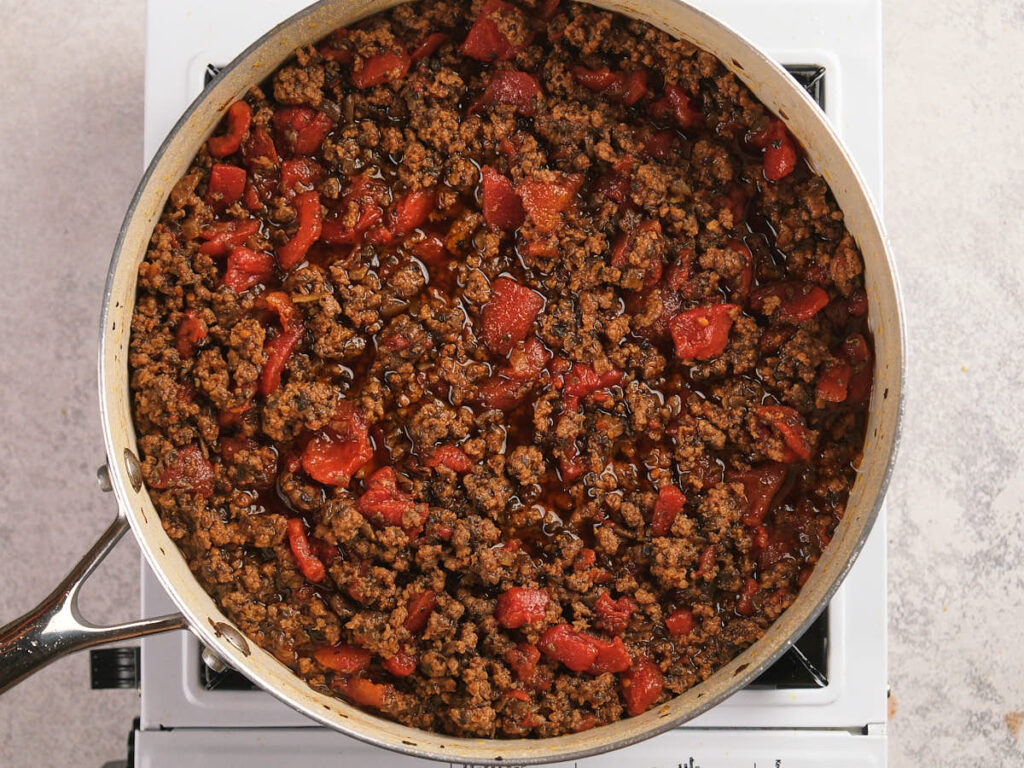 A ground beef, mushrooms and diced tomatoes are being simmered in a white skillet.