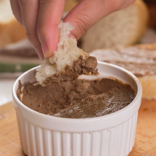 A ramekin of beef liver pate on a wooden board with a hand dipping a bread into it.