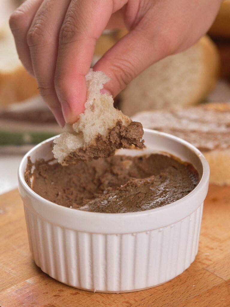 A ramekin of beef liver pate on a wooden board with a hand dipping a bread into it.
