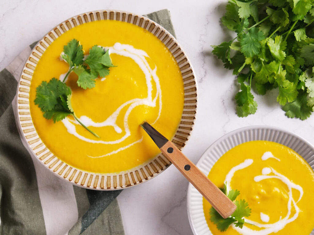 Two bowls of creamy yellow soup garnished with cilantro and a swirl of coconut milk, with fresh cilantro on the side.