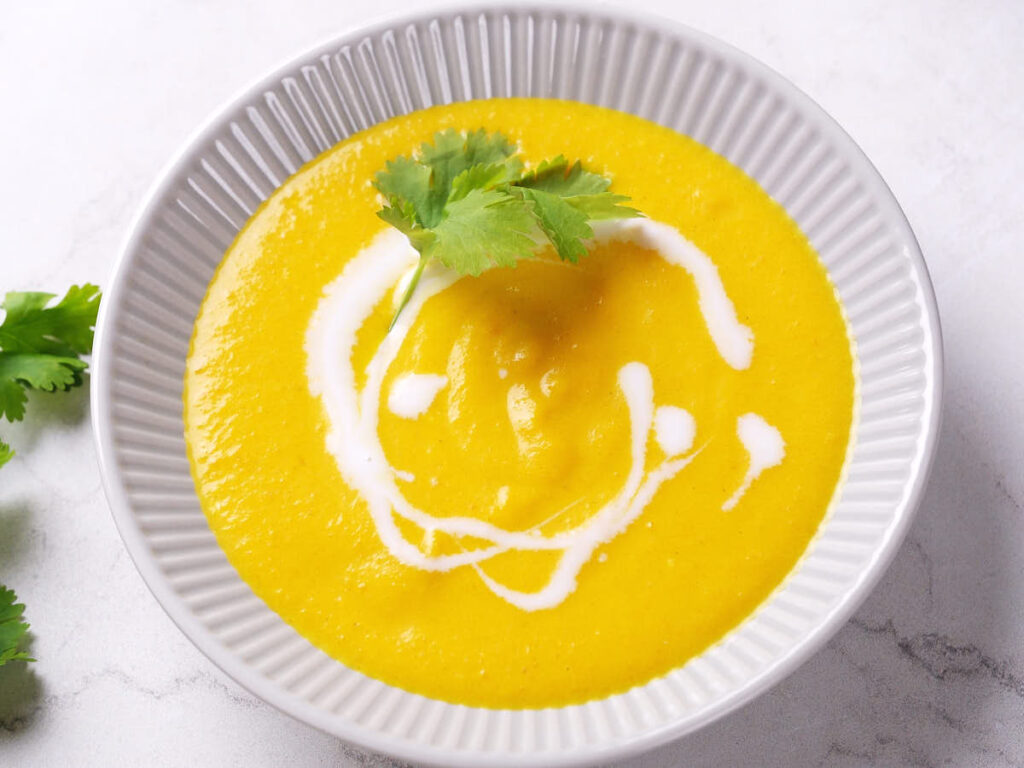 A bowl of yellow soup, garnished with a swirl of coconut milk and a sprig of cilantro.
