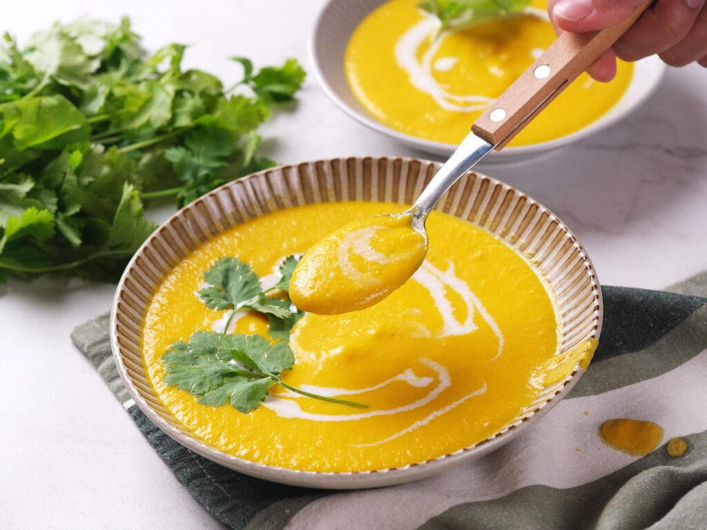 A bowl of creamy yellow soup garnished with cilantro and a swirl of coconut milk, with a spoon lifting some soup.
