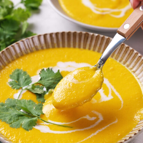 A bowl of creamy yellow soup garnished with fresh cilantro and a swirl of coconut milk, with a spoon lifting a portion.