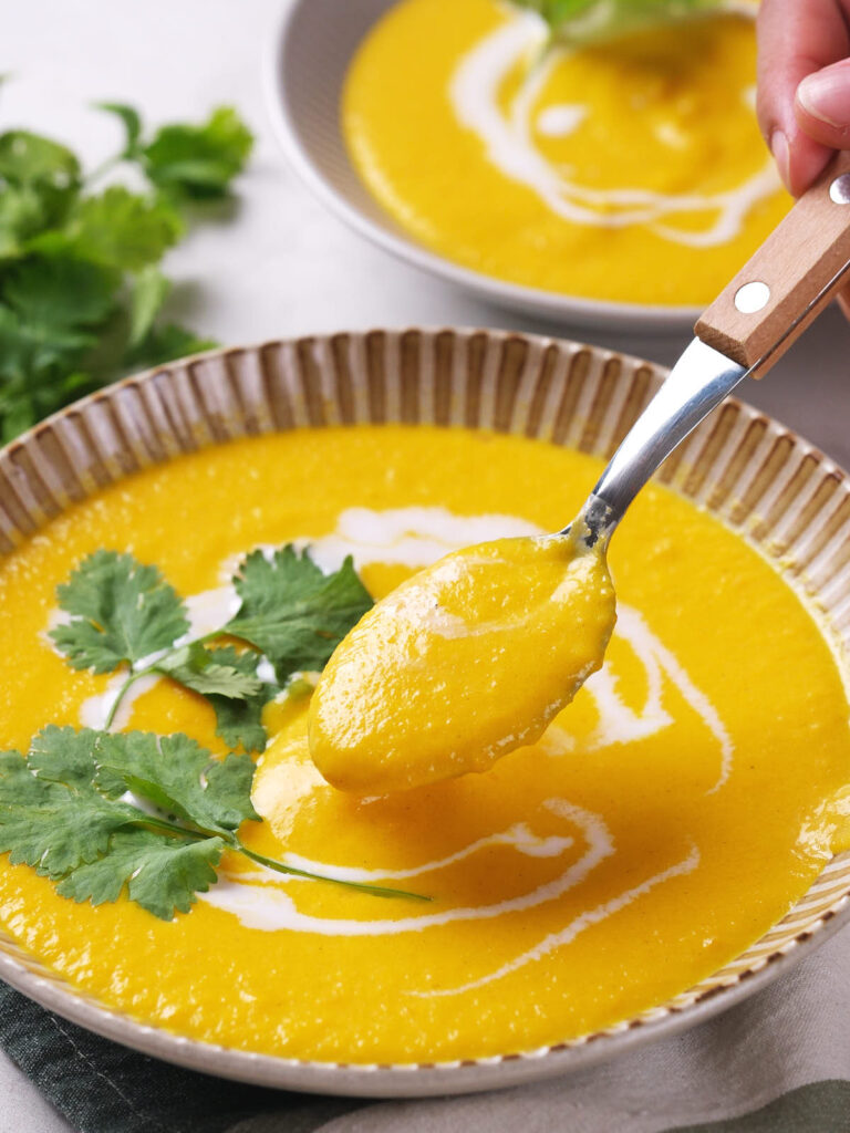 A bowl of creamy yellow soup garnished with fresh cilantro and a swirl of coconut milk, with a spoon lifting a portion.