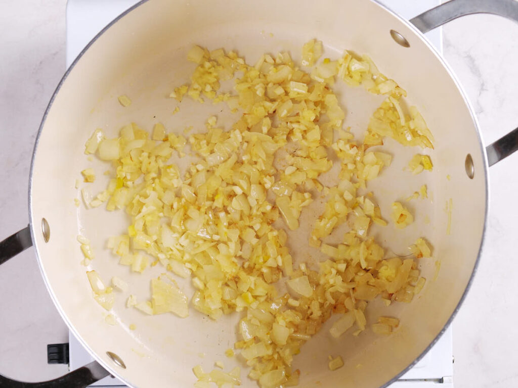 Chopped onions cooking and turning golden in a large light-colored pot on a stovetop.