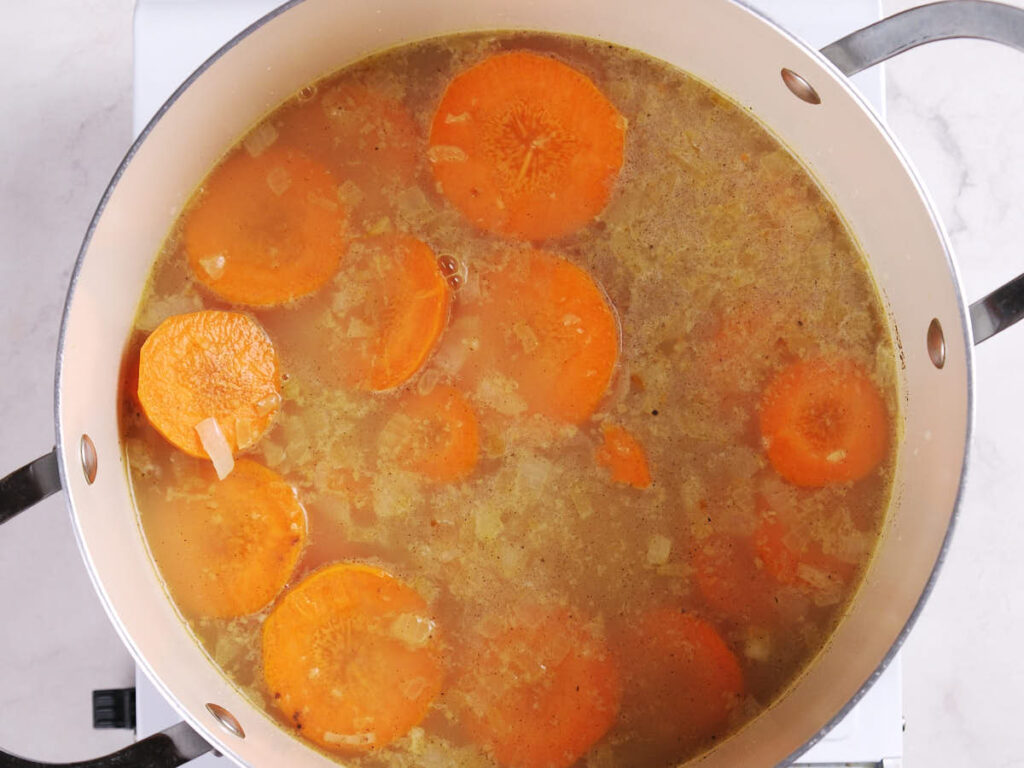 Sliced carrots simmer in broth with chopped onions in a large pot on a stovetop.