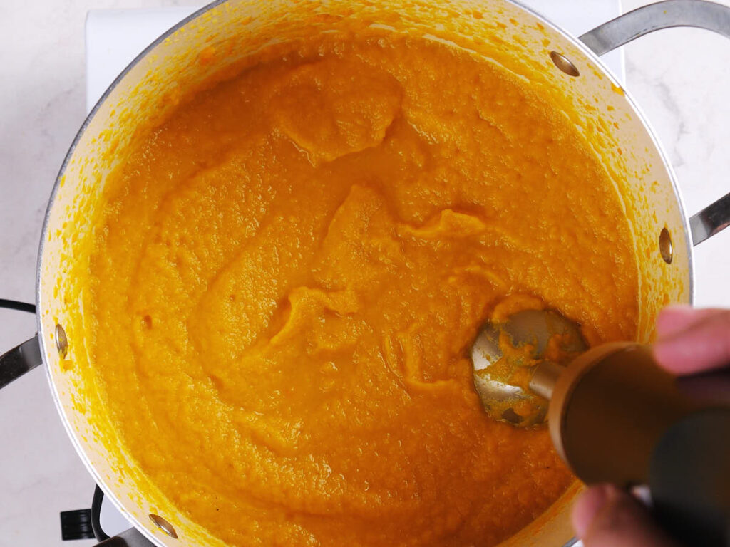 An immersion blender is being used to puree orange soup in a large pot on a white stovetop.