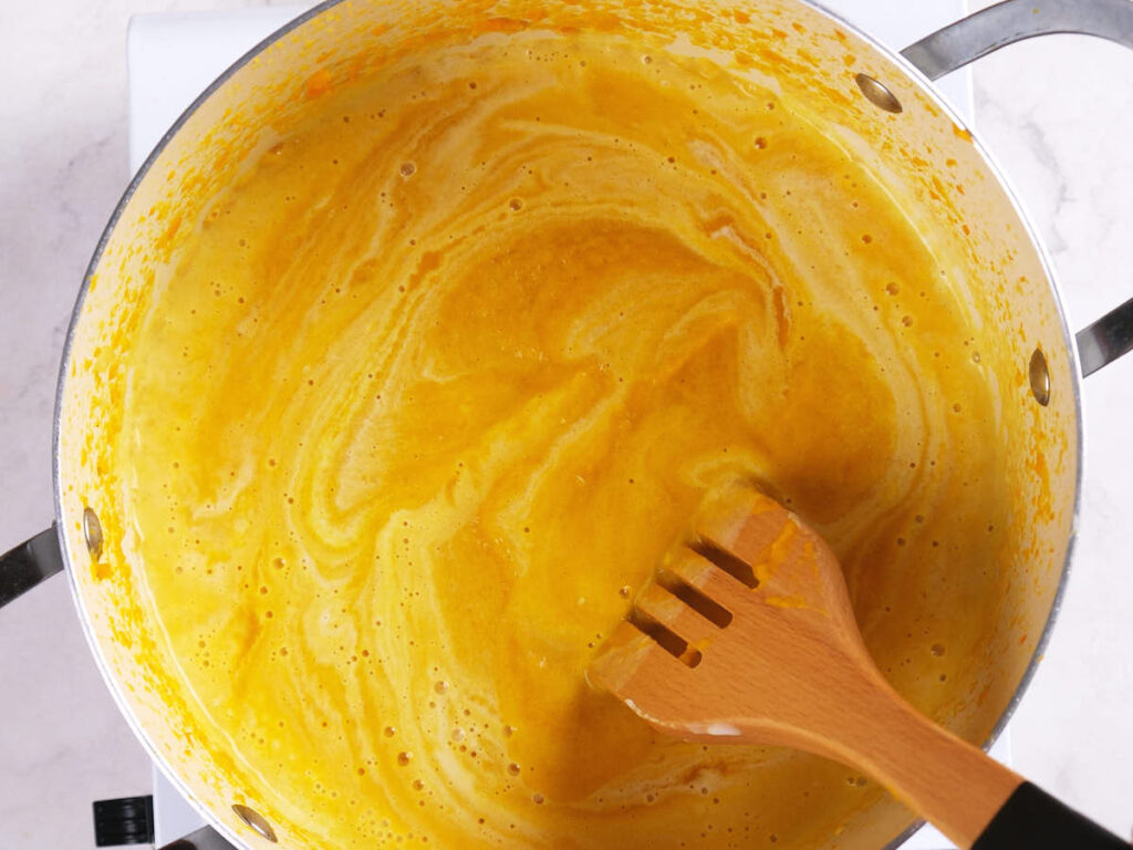 A wooden spatula stirs a yellow, creamy mixture in a large metal pot on a white surface.