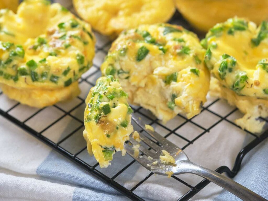 Golden muffins with chopped green herbs are cooling on a wire rack, with one partially eaten next to a fork.