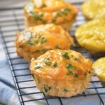 Cheesy muffin with herbs are cooling on a wire rack, underneath a table cloth.