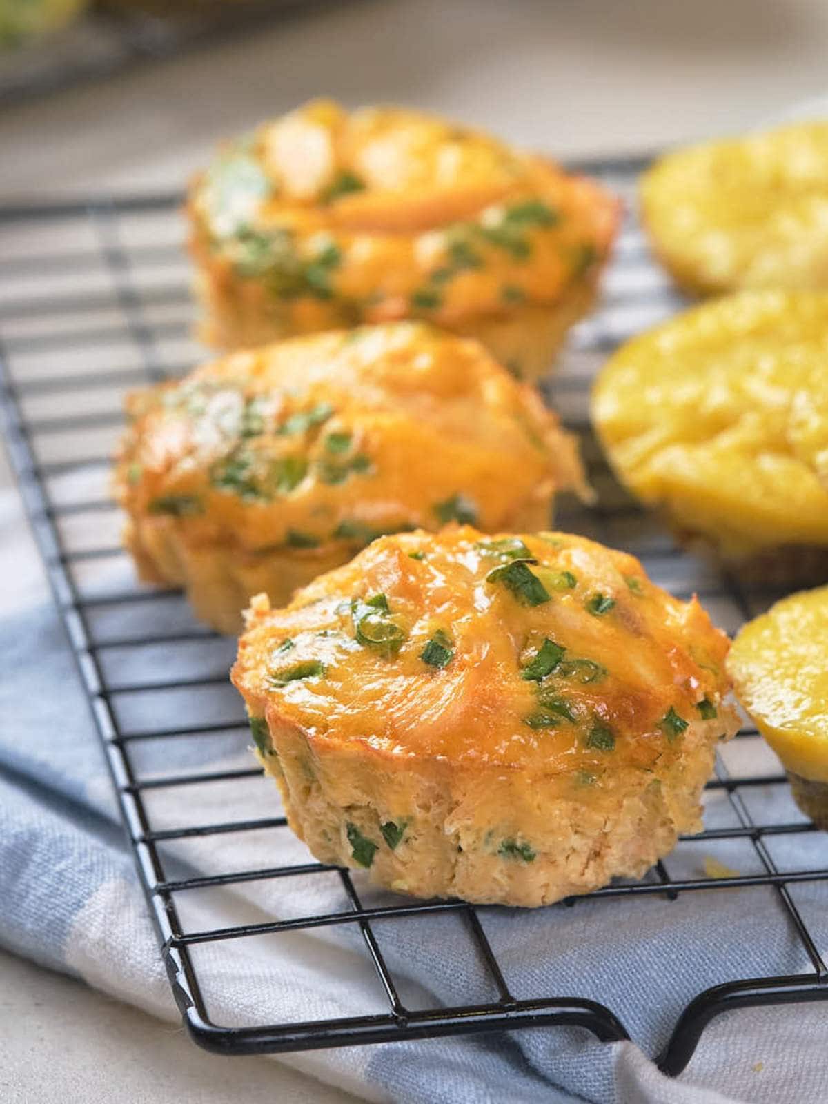Cheesy muffin with herbs are cooling on a wire rack, underneath a table cloth.