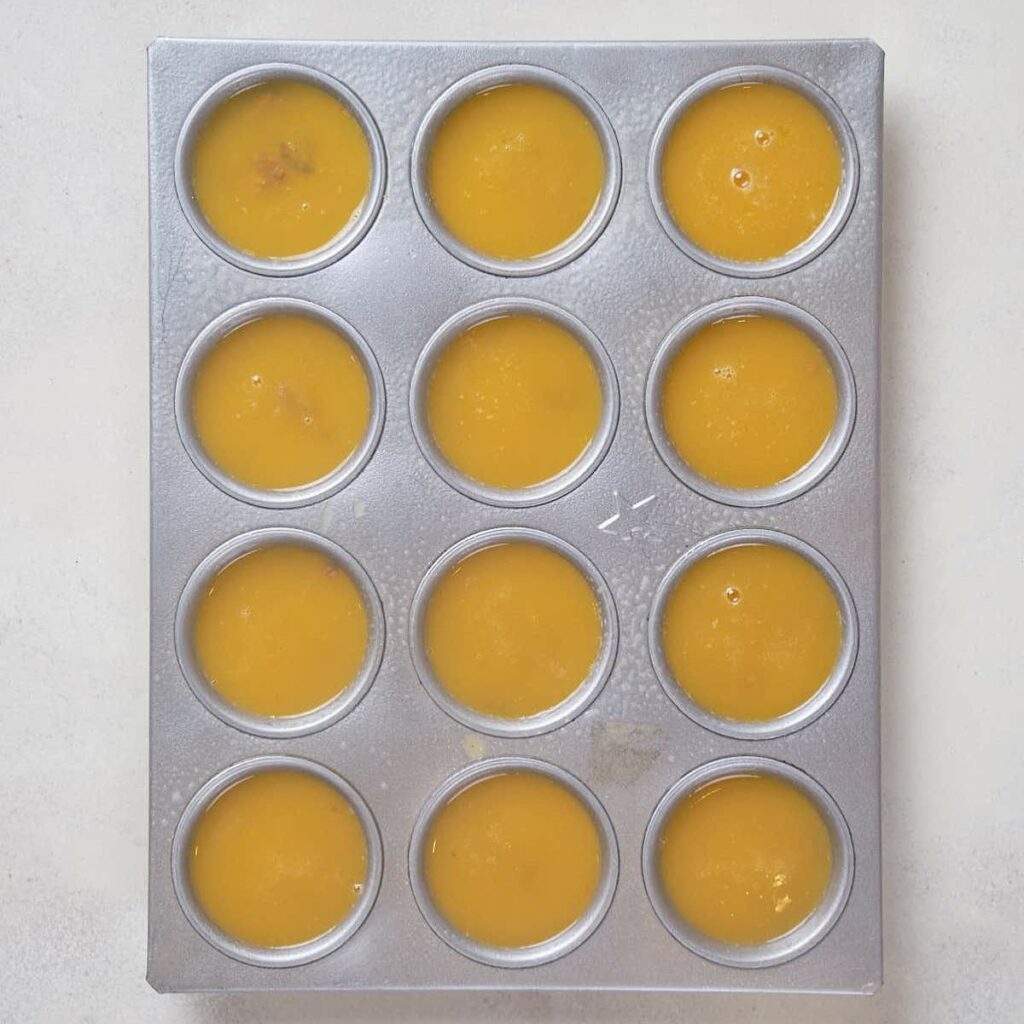 A muffin tins filled with whisked eggs and some add-ins on a white surface.