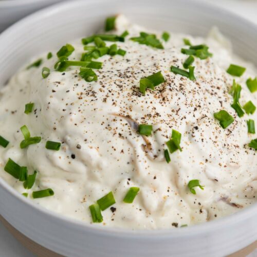 A bowl of creamy onion dip topped with chopped chives and black pepper, with chips in a separate bowl in the background.