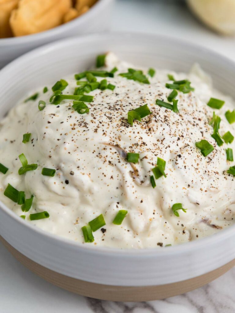 A bowl of creamy onion dip topped with chopped chives and black pepper, with chips in a separate bowl in the background.