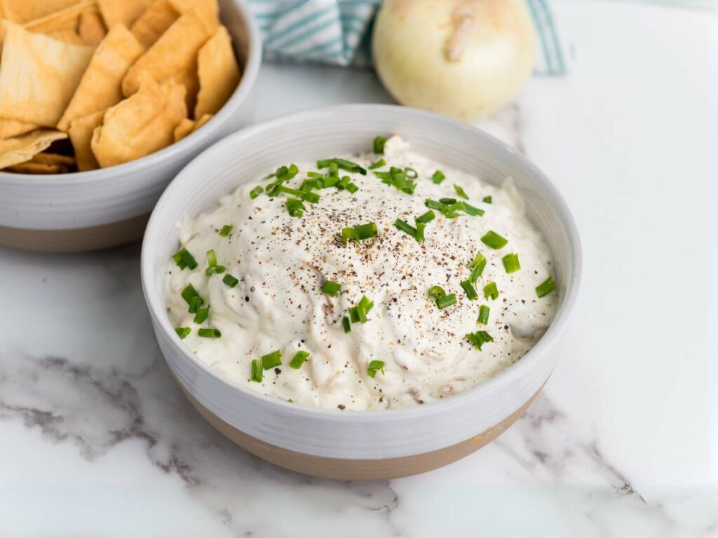 A bowl of creamy dip topped with chopped chives and black pepper sits on a marble surface beside a bowl of pita chips and a whole onion.