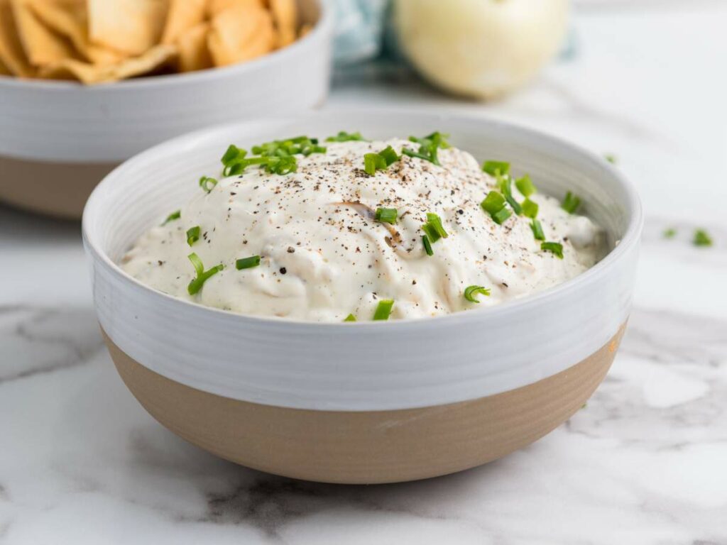 A bowl of creamy dip topped with chopped green onions and ground black pepper, with a bowl of chips in the background.