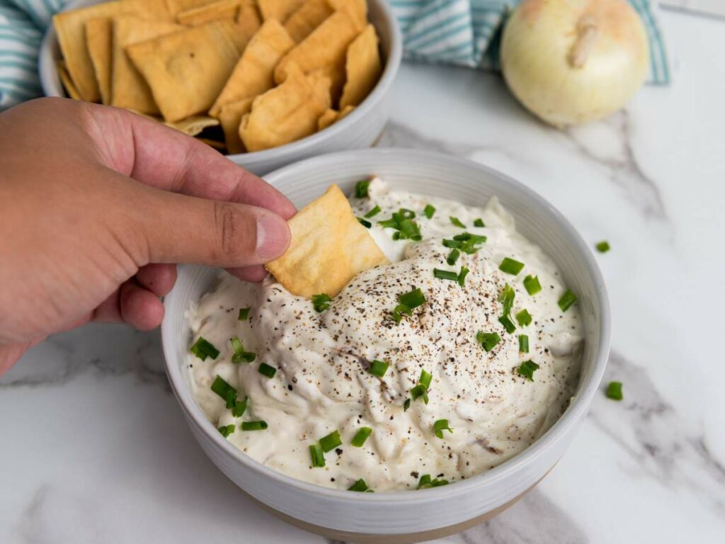 A hand dips a cracker into a bowl of onion dip topped with chopped chives and black pepper.