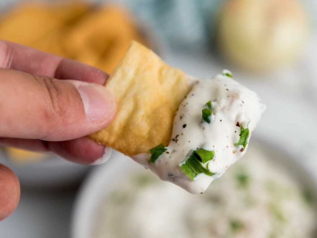 A hand holding a cracker dipped in creamy white dip garnished with chopped green onions.