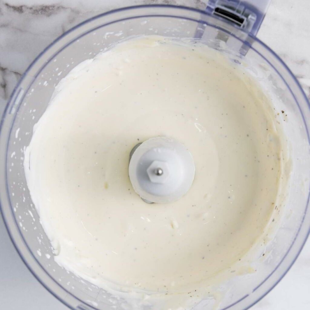 Overhead view of a food processor bowl containing a smooth, creamy dressing on a marble surface.