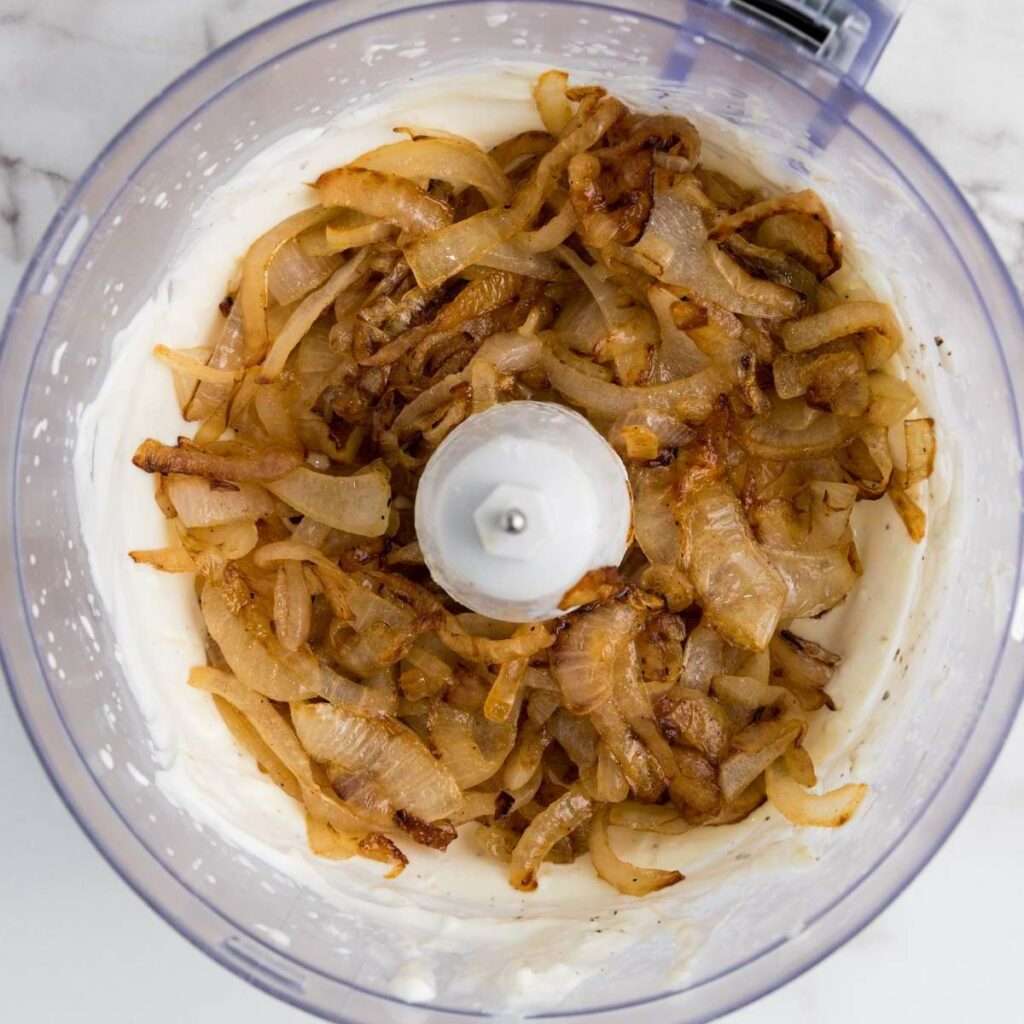 Sliced caramelized onions on top of a creamy white mixture in a food processor bowl.