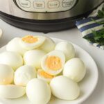 A plate of peeled hard-boiled eggs sits in front of an electric pressure cooker with fresh parsley nearby.