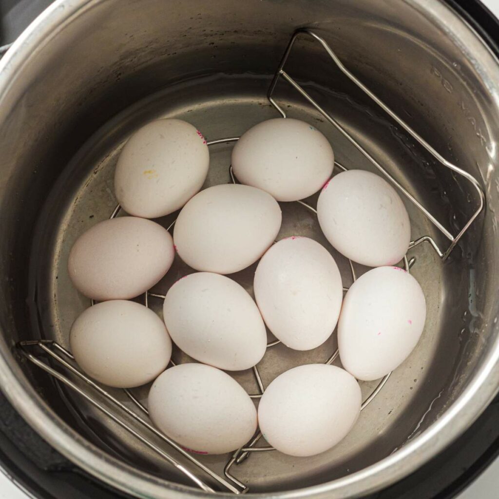 Eleven white eggs are arranged on a metal rack inside an instant pot with water at the bottom.