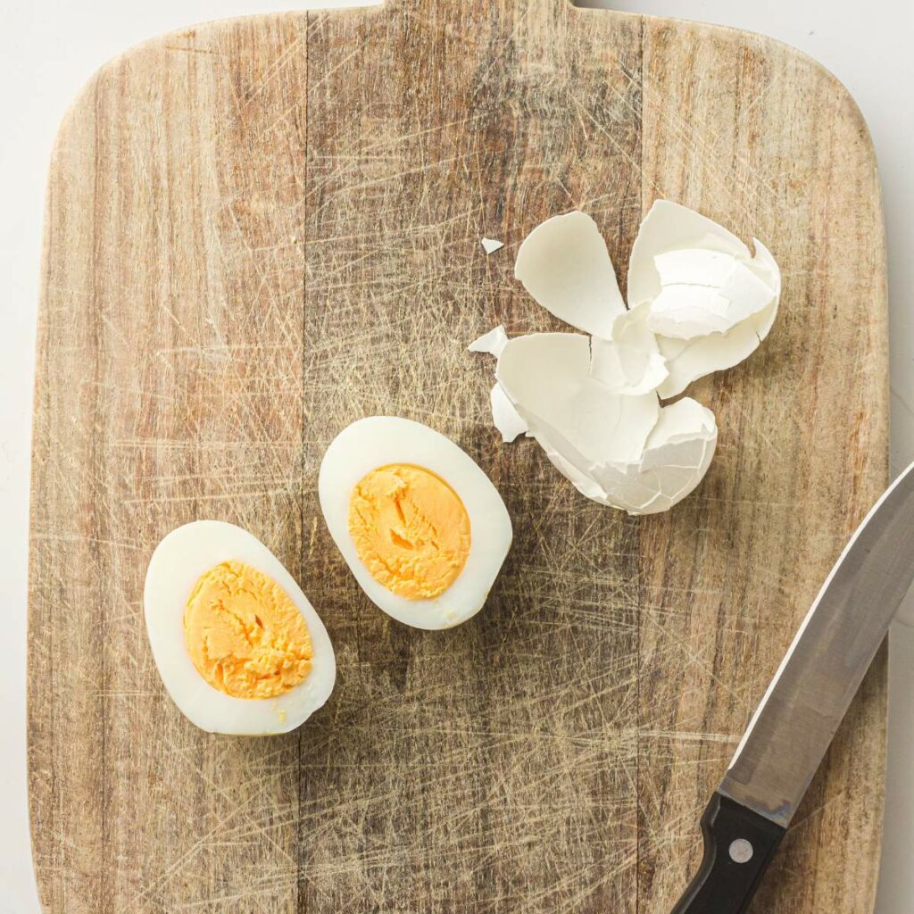 A hard-boiled egg cut in half, eggshell fragments, and a knife are on a wooden cutting board.