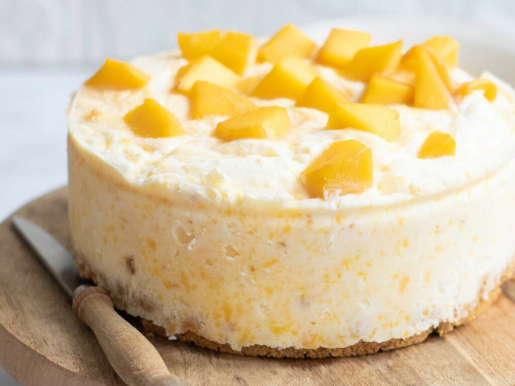 A cheesecake topped with cubes of fresh mango sits on a wooden board next to a knife.