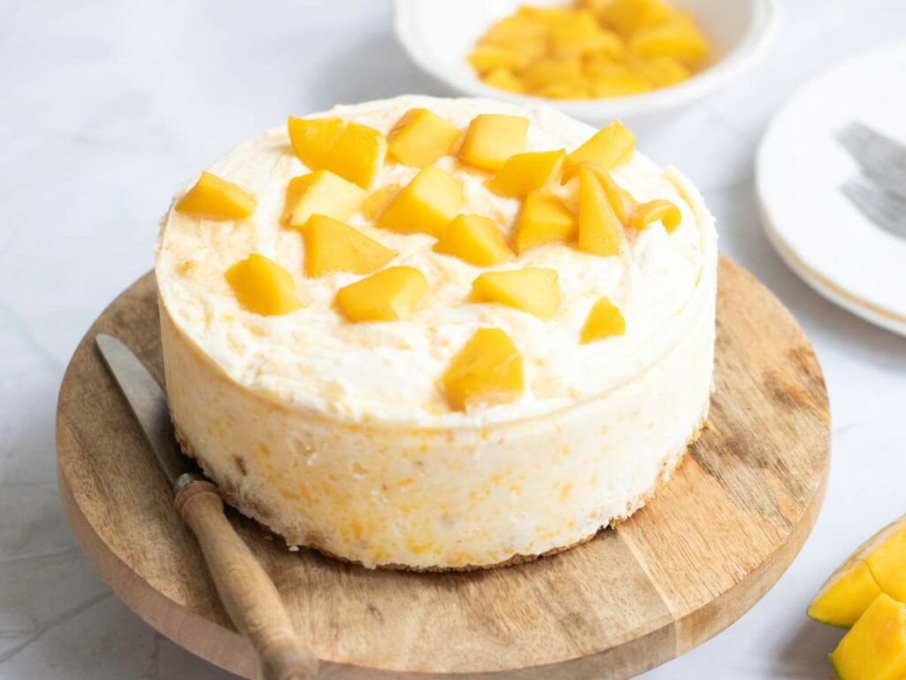 A round cheesecake topped with diced mango sits on a wooden board and a bowl of more mango pieces and a plate are in the background.