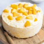 A round cheesecake topped with mango chunks, placed on a wooden board with a knife beside it.