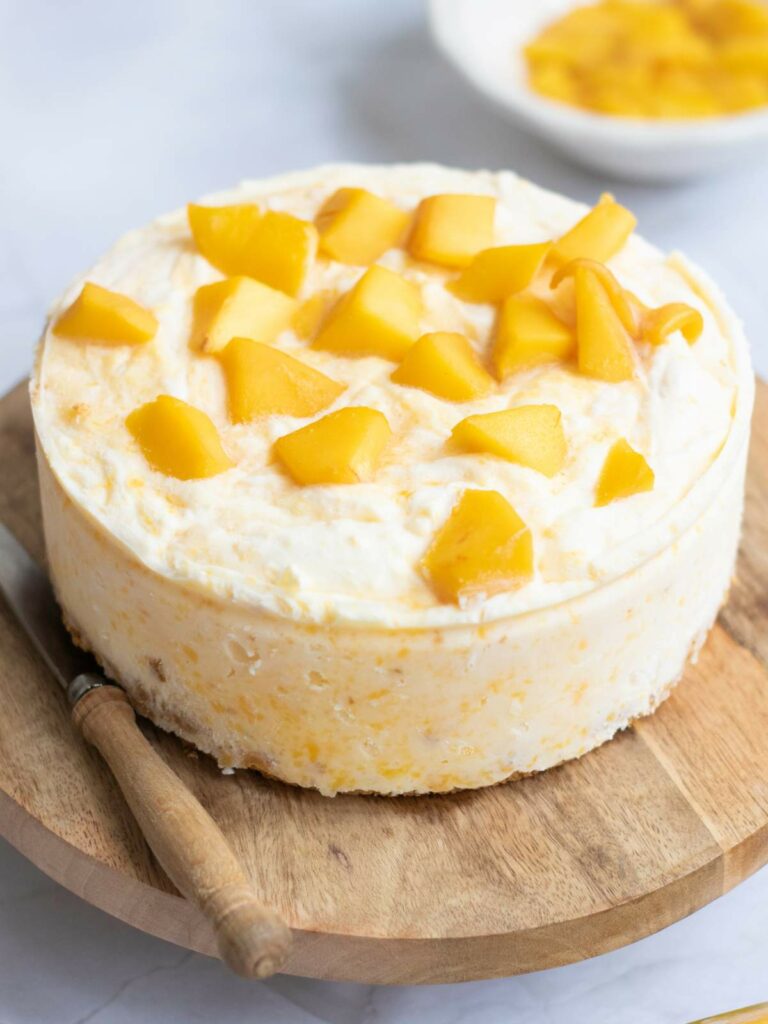 A round cheesecake topped with mango chunks, placed on a wooden board with a knife beside it.